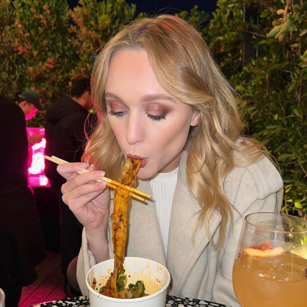 A woman with blonde hair eating long noodles with chopsticks from a white bowl at Bang Bang Noodles.