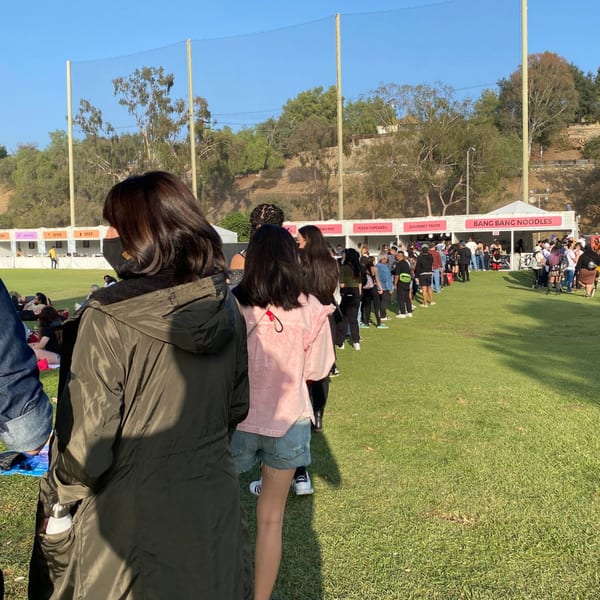 A long line of people waiting for Bang Bang Noodles at an outdoor event on a sunny day.