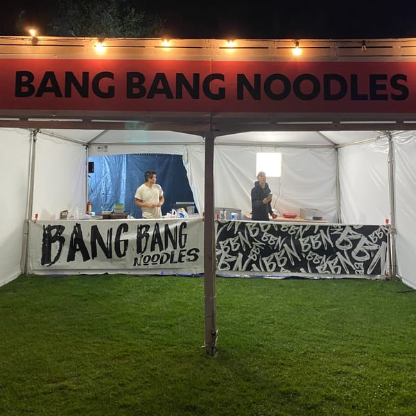 Two chefs working inside a Bang Bang Noodles tent at night, with a red sign above.