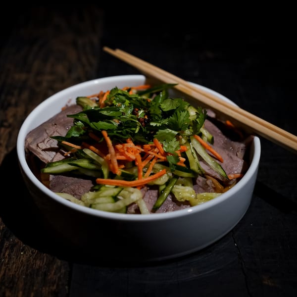 A white bowl of Bang Bang Noodles with beef, shredded carrots, cucumbers, and cilantro, with chopsticks.