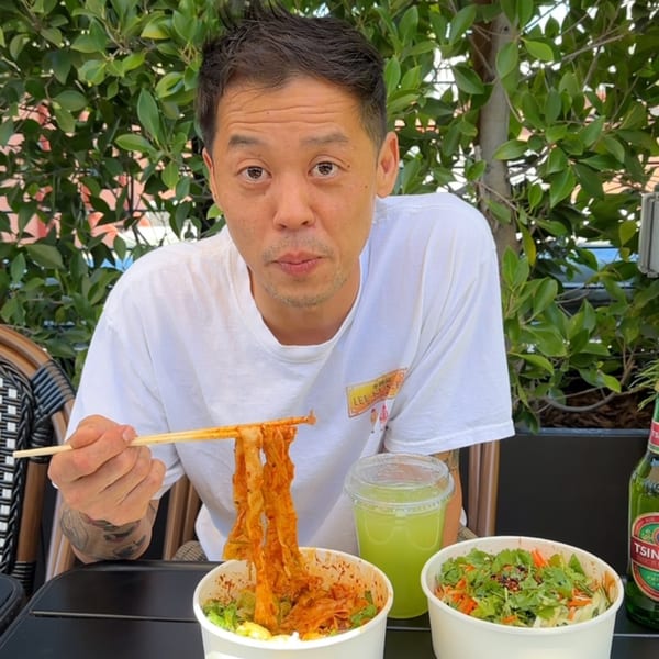 Man eating Bang Bang Noodles with chopsticks, a side salad, and a green drink outdoors.
