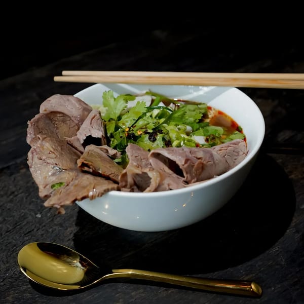 A bowl of Bang Bang Noodles with sliced beef, cilantro, and chili oil, with chopsticks and a spoon.