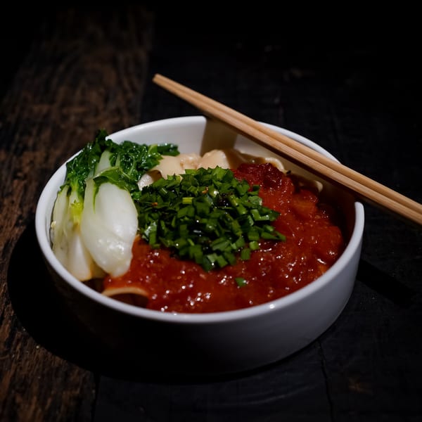 A bowl of Bang Bang Noodles with bok choy, red sauce, and green onions, with chopsticks.