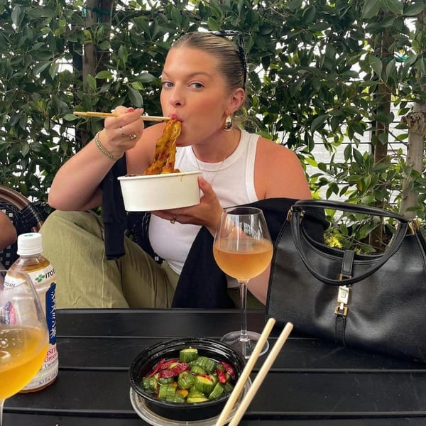 A woman eating Bang Bang Noodles with chopsticks, with a drink and side dish on a table.