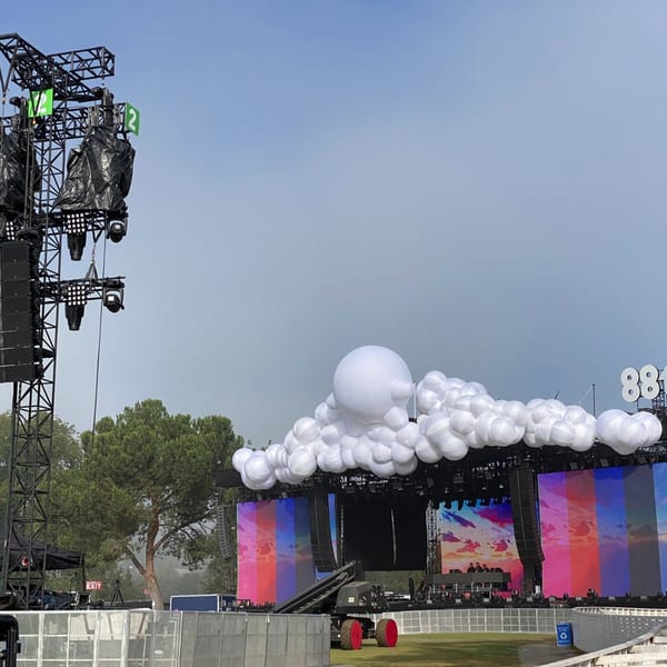 A large outdoor stage with white cloud-like decorations, large screens, and speaker towers under a cloudy sky.
