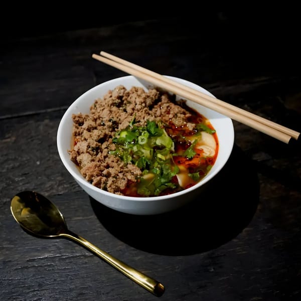 A bowl of Bang Bang Noodles with minced meat, green onions, and chili oil, with chopsticks and a spoon.